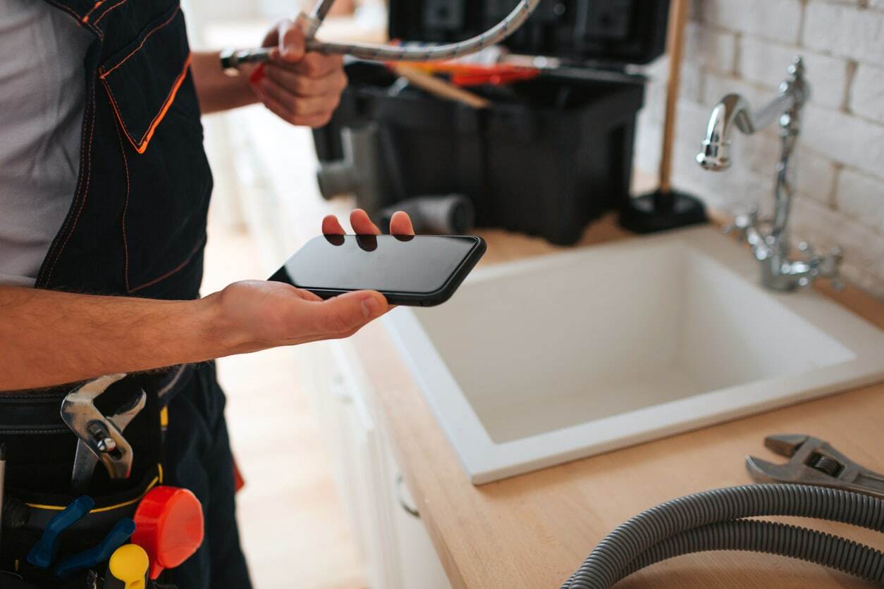 Man standing in the kitchen at sink, holding a phone and wrench 