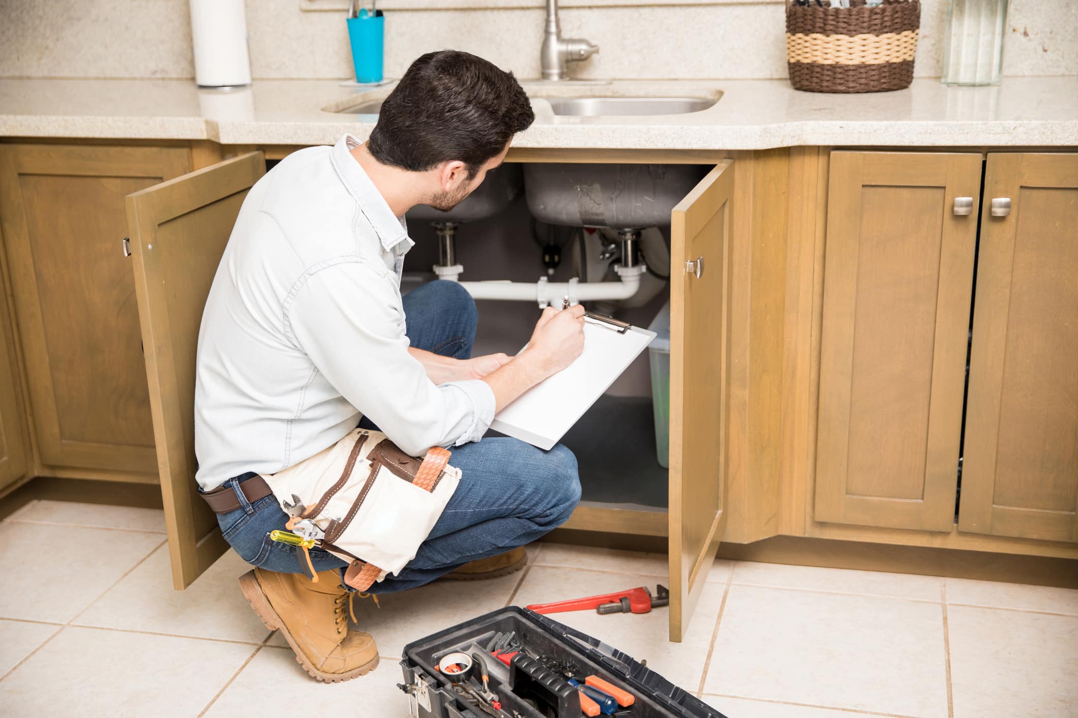 Plumber with a clipboard checking under a sink for a pipe leak